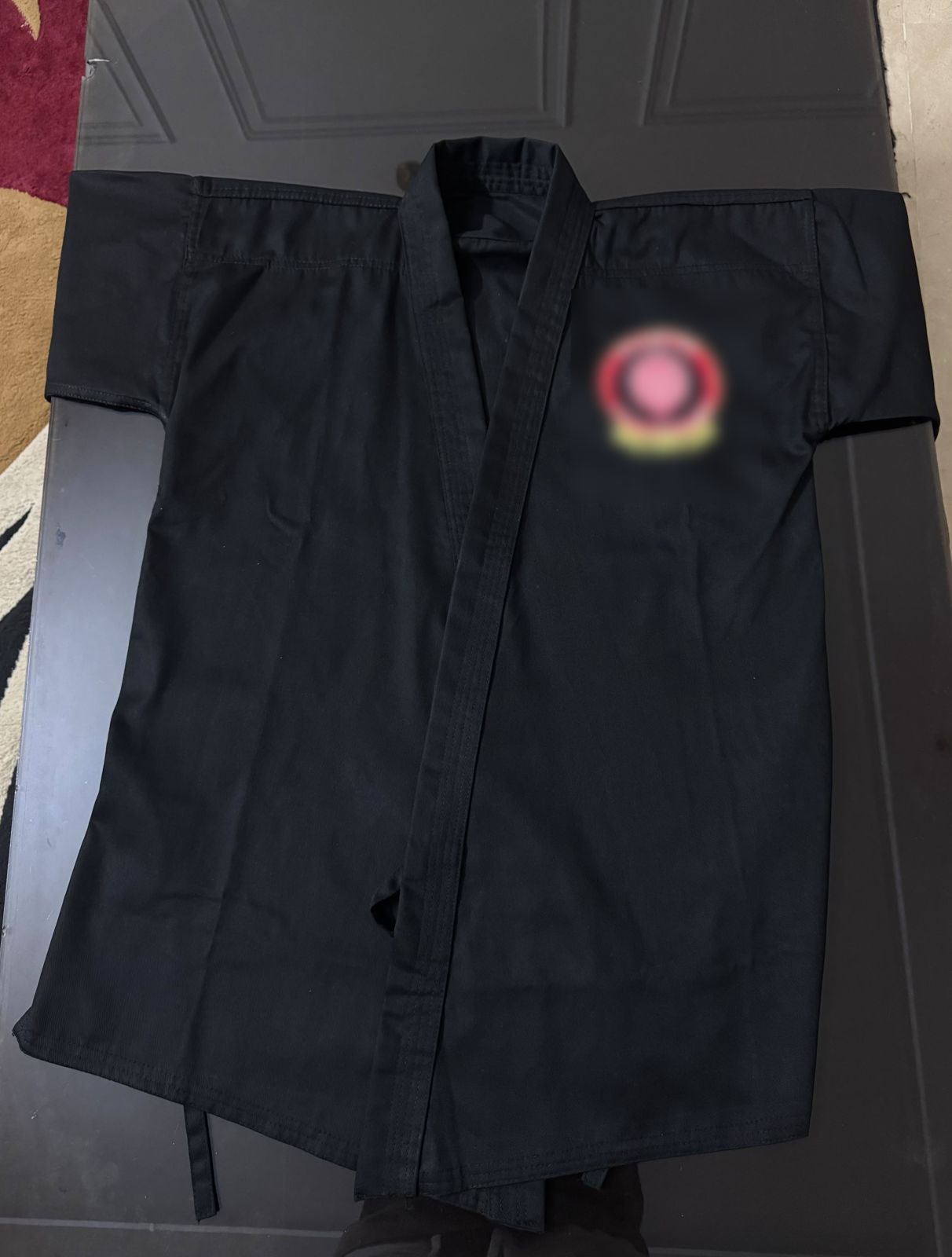 Black martial arts uniform jacket laid flat, made for a school (school logo blurred)